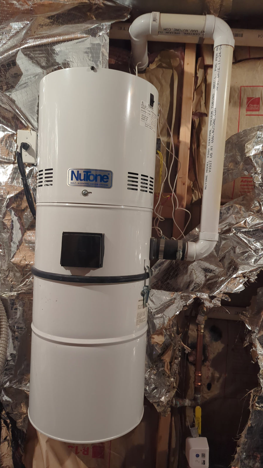 NuTone central vacuum power unit connected to white PVC piping beside insulated ductwork at AceVacuums